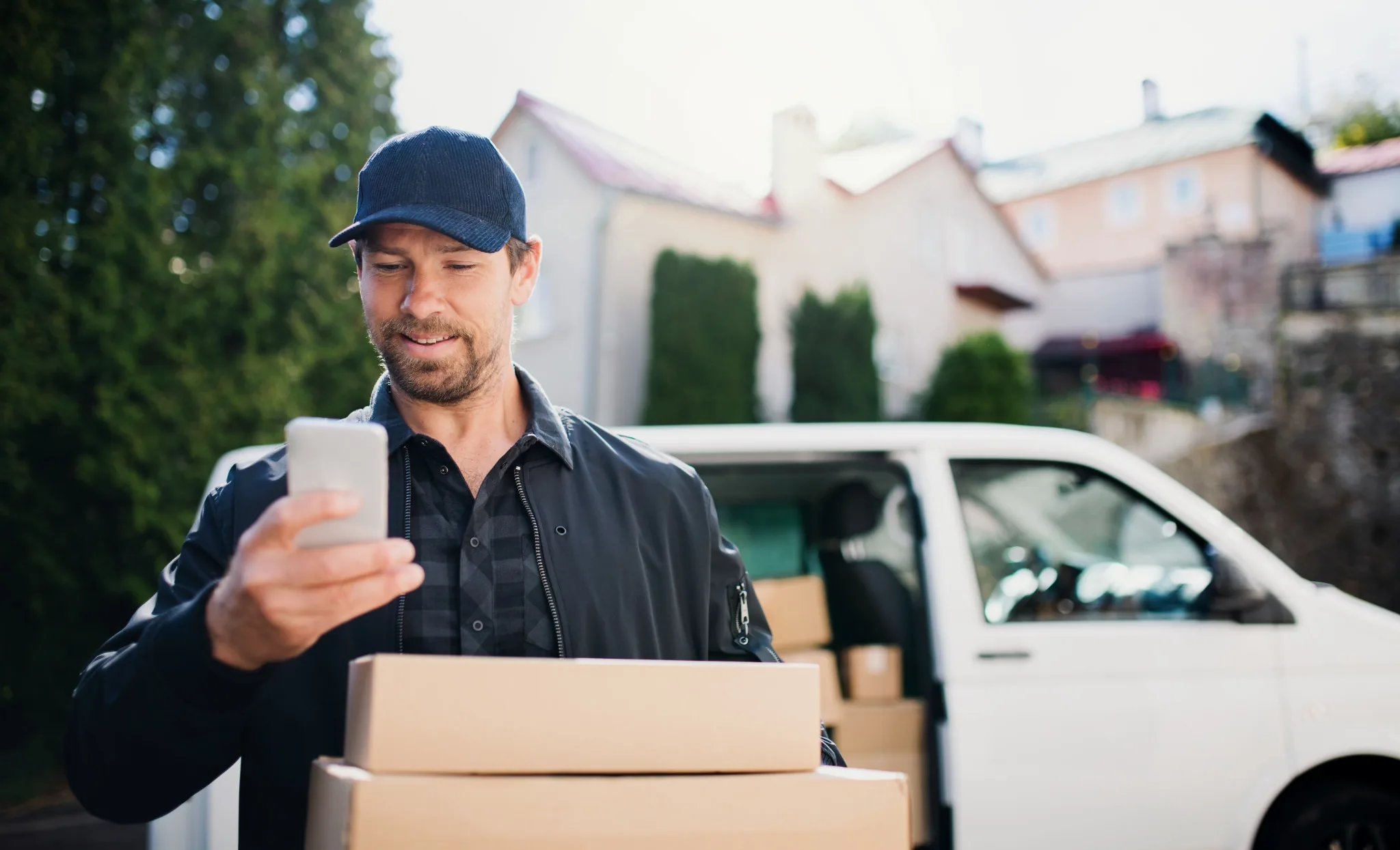 Delivery driver checking his phone with parcels in his arms beside his van